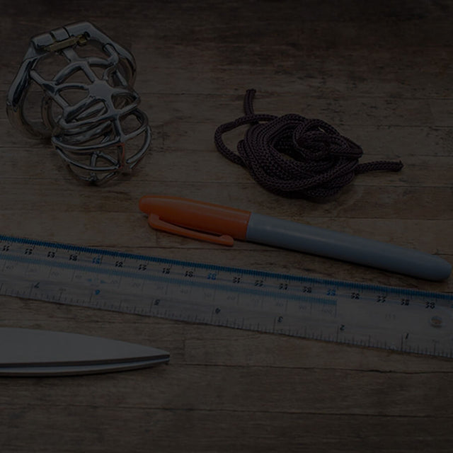 Steel chastity cage, black rope, marker pen, ruler and pair of scissors sat on a wooden board.